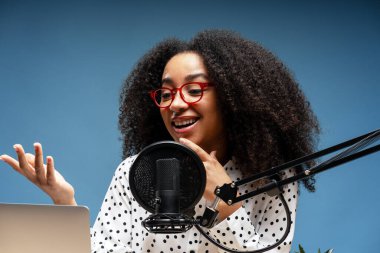 Ateşli podcast sunucusu kıvırcık saçlı ve kırmızı gözlüklü profesyonel stüdyo ortamında mikrofonlu canlı bir bölüm kaydediyor.