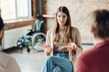Bir destek grubu toplantısında el kol hareketi yapan genç kadın psikolog, engelli bireylere zorluklarla başa çıkmaları ve zihinsel sağlığı kapsayıcı bir ortamda geliştirmeleri için rehberlik ediyor. Destek konsepti