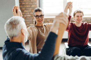 Tekerlekli sandalye kullanan kişiler de dahil olmak üzere iş arkadaşları bir daire içinde el ele tutuşuyor, birlik, işbirliği ve çeşitli ve kapsayıcı bir işyerindeki başarının kutlanmasını gösteriyorlar.