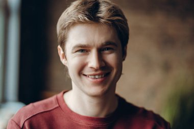 Sarı saçlı ve mavi gözlü neşeli bir gencin portresi, gündelik bir süveter giyiyor, geniş bir şekilde gülümsüyor, neşe ve pozitiflik hissi veriyor. Reklam kavramı