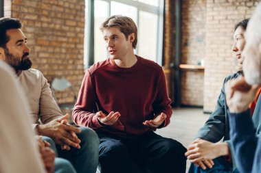 Genç bir adam grup terapisi sırasında kişisel mücadelelerini açıyor ve paylaşıyor, katılımcılar arasında iyileşme ve büyüme için destekleyici ve anlayışlı bir atmosfer sağlıyor