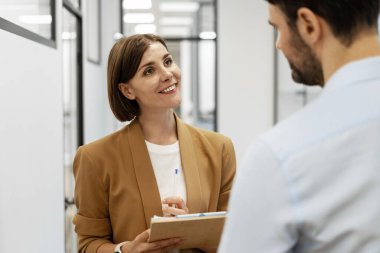 İş kadını elinde bir pano tutarken ve bir ofis koridorunda meslektaşıyla tartışırken gülümserken takım çalışmasını ve işbirliğini teşvik ediyor.