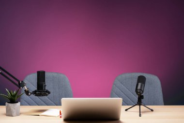 Canlı pembe ışıkla aydınlatılan mikrofonlar, dizüstü bilgisayarlar ve sandalyelerle modern podcast stüdyosu, ses içeriği oluşturulması için profesyonel ve davetkar bir atmosfer yaratıyor.