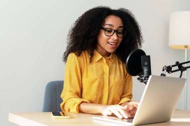 Gülümseyen Afro-Amerikan kadın dizüstü bilgisayar ve mikrofon kullanarak podcast yapıyor. Canlı, canlı konsept