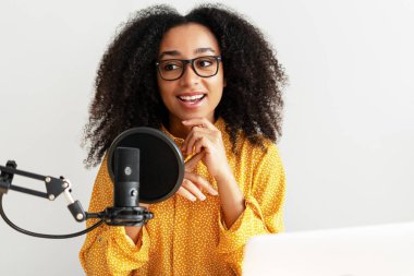 Gülümseyen genç bir kadın podcaster ev stüdyo ortamında podcast 'ini kaydederken mikrofona konuşuyor, notlar ve kurgu için dizüstü bilgisayar kullanıyor. Çevrimiçi, canlı, canlı konsept