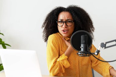 Üzgün, mutsuz, gözlüklü Afrikalı Amerikalı kadın podcast 'i kaydediyor, laptop kullanıyor ve stüdyoda oturuyor. Çevrimiçi teknoloji kavramı