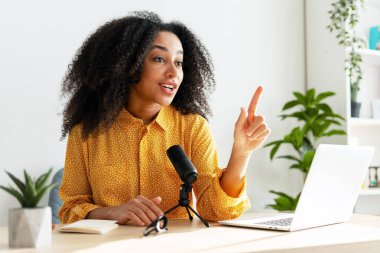 Güzel Afro-Amerikan içerik yaratıcısı şık sarı gömlekli podcast kaydediyor ve parmağıyla işaret ediyor. Çevrimiçi teknoloji kavramı