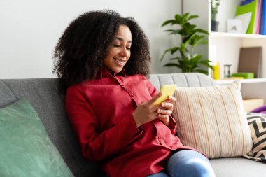 Oturma odasındaki rahat koltukta dinlenen gülümseyen Afro-Amerikalı kadın cep telefonunu kullanıyor ve evde boş zamanlarının tadını çıkarıyor, sosyal medyaya dalmış. Çevrimiçi teknoloji kavramı