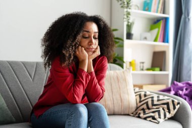 Kıvırcık saçlı, günlük giysiler içinde üzgün bir kadın kanepeye oturmuş elleri üzerinde dinleniyor, düşüncelere dalmış, rahat oturma odasında yalnızlık ve depresyon yaşıyor.