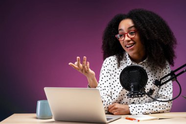 Genç bir kadın bir stüdyoda dizüstü bilgisayar ve profesyonel bir mikrofon kullanarak podcast yaparken konuşuyor ve el kol hareketi yapıyor.