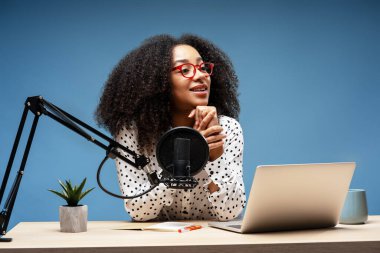 Kıvırcık saçlı ve kırmızı gözlüklü genç bir kadın, mikrofon ve dizüstü bilgisayarla masada oturuyor, mavi arka planı olan bir stüdyoda podcast kaydediyor, ellerini sıkarak yüzünü çeviriyor. Çevrimiçi teknoloji kavramı