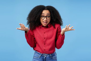 Kafası karışmış, kıvırcık saçlı ve gözlüklü, omuzlarını silkeleyen ve avuçlarını mavi bir arka plana doğru kaldıran Afro-Amerikan bir kadının stüdyo portresi.