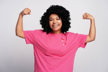 Gülümseyen kadın güç gösteriyor ve kaslarını kasıyor pembe kurdeleli pembe tişört giyiyor, göğüs kanseri bilinçlendirme kampanyasını destekliyor.