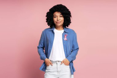 Göğüs kanserini destekleyen pembe kurdeleli, pembe arka planda poz veren genç bir kadının stüdyo resmi.