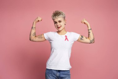 Beyaz tişörtüne pembe kurdele takmış kaslarını esneten, göğüs kanseri farkındalığını destekleyen, pembe arka planda izole edilmiş bir kadın.