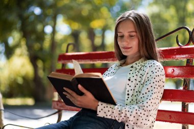 Öğrenci, güneşli bir günde yeşil bir parkta kırmızı bir bankta oturmuş kitap okumaktan zevk alıyor.