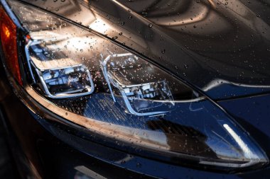 Siyah bir arabanın şık farları su damlacıklarıyla kaplıdır. Çevredeki ışık kaynaklarını yansıtarak soyut bir model oluşturur.