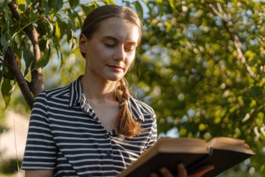 Bahçede oturan bir kadın, bir kitap tutuyor ve açık havada huzurlu bir anın tadını çıkarıyor, yemyeşil ve sıcak güneş ışığıyla çevrili.
