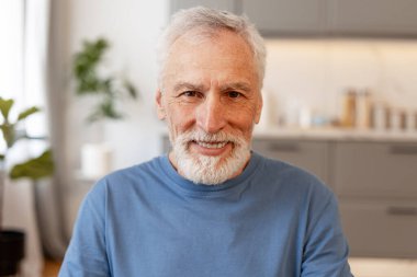 Gri saçlı ve sakallı mutlu emekli adam modern ve parlak evinde otururken gülümsüyor emekliliğinin ve sağlıklı yaşam tarzının tadını çıkarıyor. Reklam kavramı