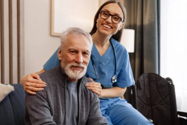 Genç hemşire gülümseyip, evde ya da huzurevinde, sağlık hizmetleri ve tıbbi yardım konseptinde oturan yaşlı ve mutlu bir adamı destekliyor. Yardımcı kavram