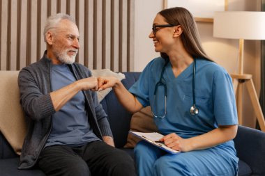 Gülümseyen kadın doktor ve mutlu kıdemli adam ev ziyareti sırasında başarılı bir muayeneden sonra yumruk tokuşturuyor, sağlık hizmetleri uzmanı ile hasta arasındaki güven ve olumlu ilişkiyi gösteriyor.