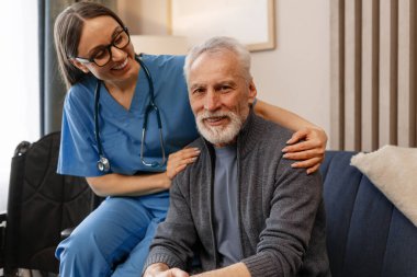 Steteskoplu genç hemşire gülümseyip, kanepede oturan, yaşlı bir hastaya evde ya da huzurevinde yardım eden yaşlı bir adamı destekliyor.