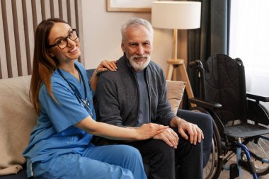 Gülümseyen hemşire, yaşlı bir adamın yanında oturuyor ve arka planda tekerlekli sandalyeyle elini tutarken onu rahatlatıyor. Hareket sorunu olabileceğini gösteriyor. Destek konsepti