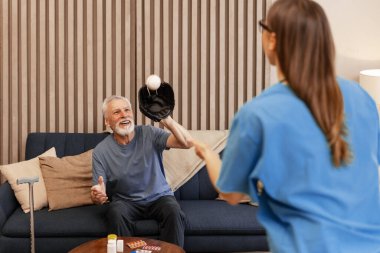Bastonlu yaşlı adam hemşiresiyle beysbol oynuyor, modern bir evin oturma odasında oyun oynuyor, sağlığını ve sağlığını geliştiriyor. Rehabilitasyon kavramı