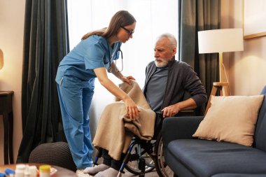 Genç hemşire yaşlı adama bakıyor, tekerlekli sandalyede otururken onu battaniyeyle örtüyor, yaşlı hastalara bakım ve destek sağlıyor. Destek konsepti