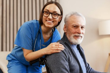 Genç hemşire tekerlekli sandalyede oturan mutlu yaşlı adamı rahatlatıyor, sağlık görevlisi huzurevinde emekli bir hastaya yardım ediyor, ev bakımı konsepti