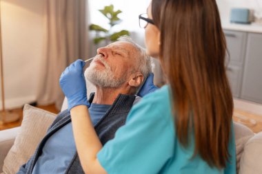 Eldiven giyen genç hemşire, oturma odasındaki koltukta rahat oturan yaşlı bir adama genizden örnek alıp salgın sırasında gerekli ev sağlığı hizmetlerini sağlıyor.