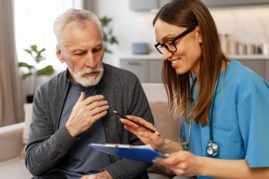 Mavi önlük ve steteskop takmış gülümseyen kadın doktor elinde pano ve kalemle evdeki koltukta oturan ve semptomlarını açıklayan yaşlı adamla konuşurken onu işaret ediyor.