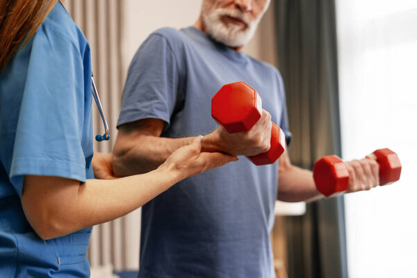 Медсестра оказывает помощь старшему мужчине в поднятии красных гантелей, подчеркивая реабилитацию и восстановление сил в благоприятной обстановке домашнего здравоохранения, повышение общего благосостояния и мобильности