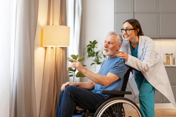 Happy senior man sitting in a wheelchair points at something through the window while a smiling nurse supports him by putting a hand on his shoulder. Support concept