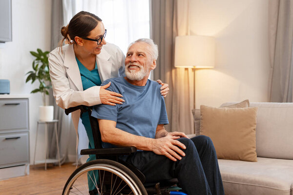 Female doctor is supporting smiling senior man sitting in wheelchair at home, they are looking at each other and having pleasant conversation. Support concept
