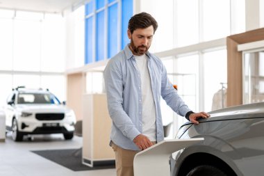 Yenilikçi modellerle dolu modern bir araba galerisinde, elektrikli bir aracın şarj limanını inceleyen sakallı bir adam.