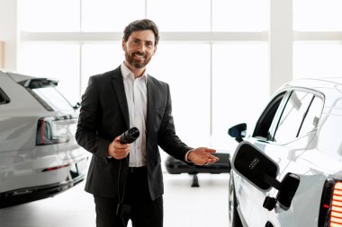 Otomobil galerisinde yeni bir elektrikli arabayı tanıtırken elinde şarj kablosu tutan satıcı, sürdürülebilir taşımacılıktaki gelişmeleri vurguluyor