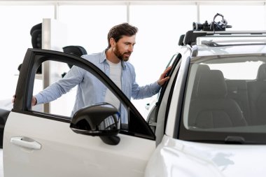 Sakallı adam, beyaz bir arabanın kapısını açıyor. Teçhizatlı bir araba galerisinin içinde tavan rafı var.