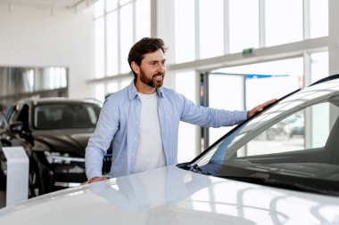 Galeride otomobil seçen, araba kaputuna dokunan, gülümseyen, yeni bir araba alan mutlu bir adam.