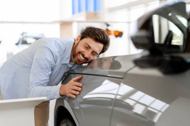 Neşeli bir adam yeni arabasını bir araba galerisinde kucaklıyor. Satın almasından duyduğu neşe ve memnuniyeti dile getiriyor.