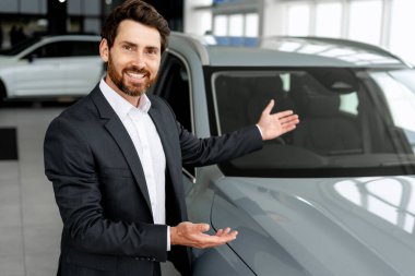 Gülümseyen araba satıcısı modern bir bayilikte yeni bir araç sunuyor. Otomotiv endüstrisinde profesyonellik ve kendine güven gösteriyor.