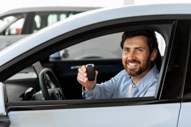 Gülümseyen adam elinde araba anahtarıyla galeride yeni bir araca binmiş başarılı bir alımı kutluyor ve mülkiyetin heyecanının tadını çıkarıyor.