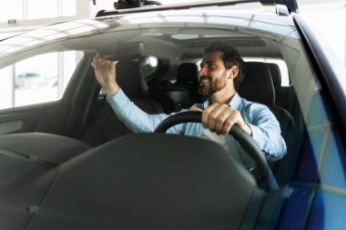 Mutlu sakallı iş adamı araba galerisinde yeni arabada otururken dikiz aynasını ayarlıyor.