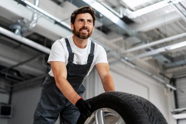 Bir oto tamirhanesinde lastik iten kendinden emin bir tamirci, başka tarafa bakarken gülümseyen, uzmanlığı ve kendini işine adamış biri.