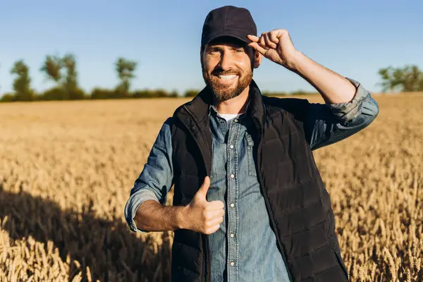 Güneşli bir yaz gününde altın buğday tarlasında dikilip hasattan memnuniyetini ifade ederken başparmağını kaldırıp şapkasına dokunan gülümseyen çiftçi portresi. Çiftçilik kavramı