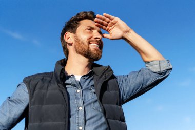 Neşeli bir çiftçinin portresi, arka planda mavi gökyüzüne bakarken gözlerini güneşten koruyor.