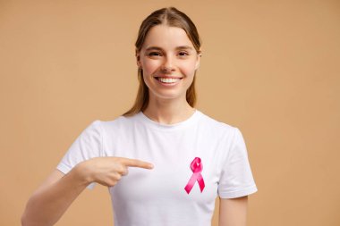 Gülümseyen kadın göğüs kanseri farkındalığı için pembe kurdeleye işaret ediyor, bej arka planda beyaz tişört giyiyor. Sağlık hizmeti kavramı