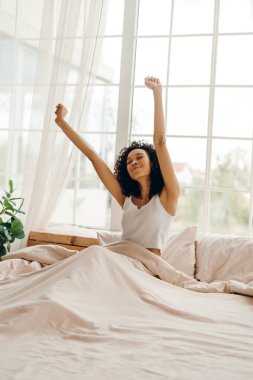 Gülümseyen Afrikalı Amerikalı genç bir kadının portresi. Gözleri kapalı, yatakta dinleniyor, kollarını evde sabah ışığında açıyor. Yaşam tarzı kavramı