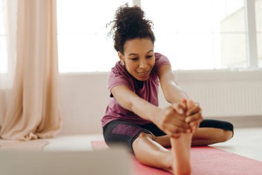 Gülümseyen Afro-Amerikan atletik giyimli aktif genç kadın evinde bacak esneme hareketi yapıyor. Spor, sağlıklı bir kavram.