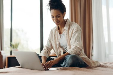 Mutlu Afrika kökenli Amerikalı kadın evinde yatağında rahat bir şekilde oturuyor dizüstü bilgisayarıyla çalışırken gülümsüyor ve ekrana bakıyor. Çevrimiçi iş kavramı